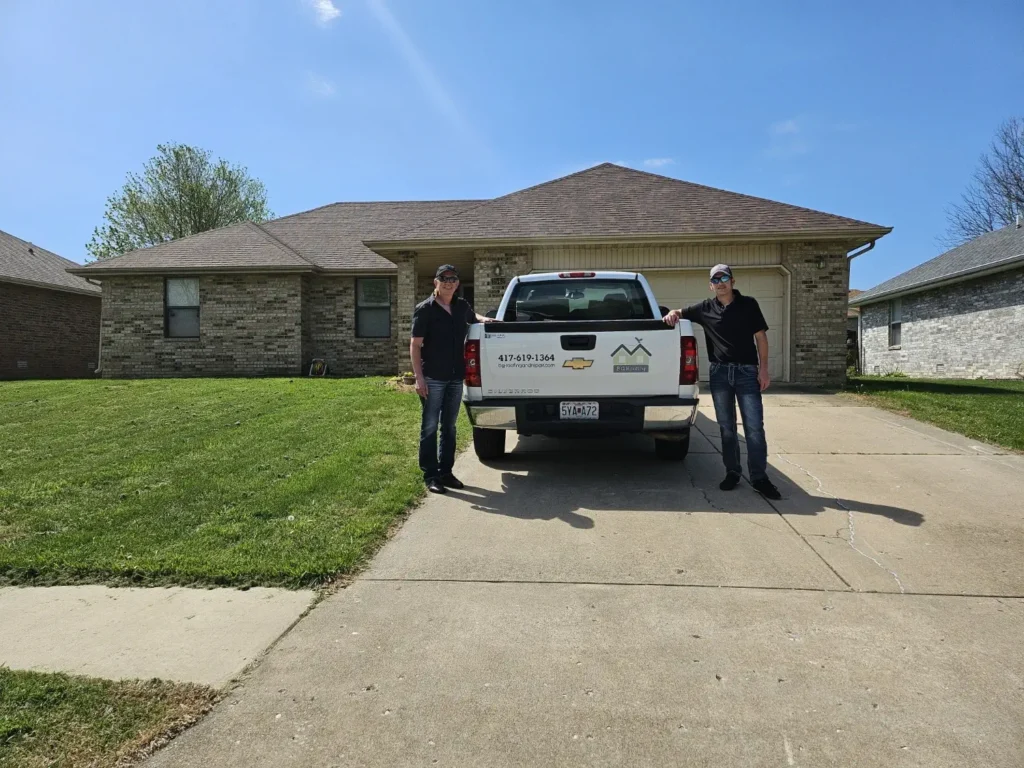 Two men standing beside a white pickup truck parked in front of a suburban brick house undergoing roof improvement on a sunny day.