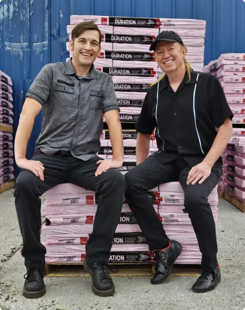 Gene and Jesse from BG Roofing smiling and sitting on a stack of roof shingles outside a warehouse.